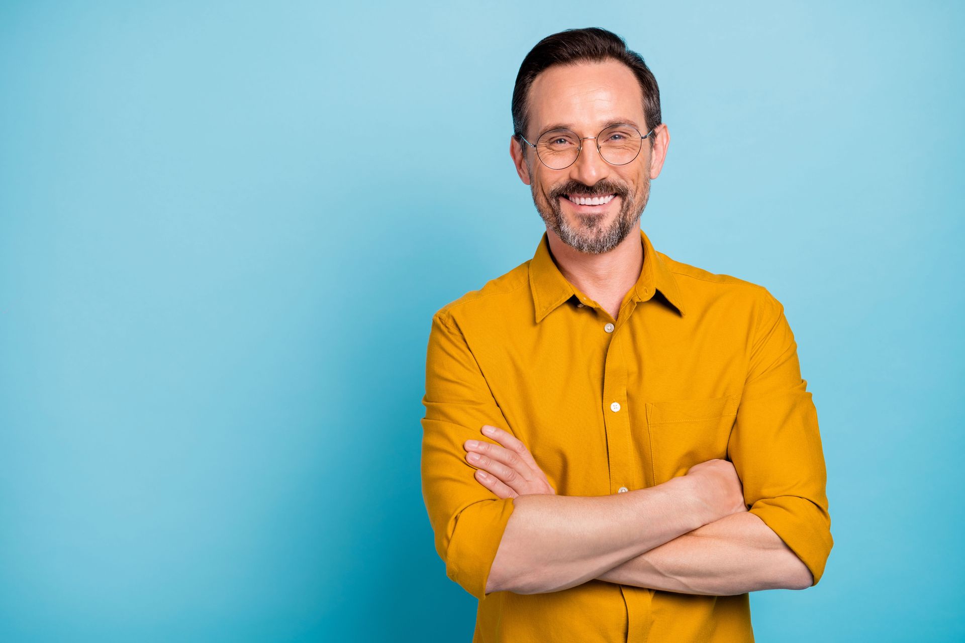 A man in a yellow shirt and glasses is standing with his arms crossed and smiling.