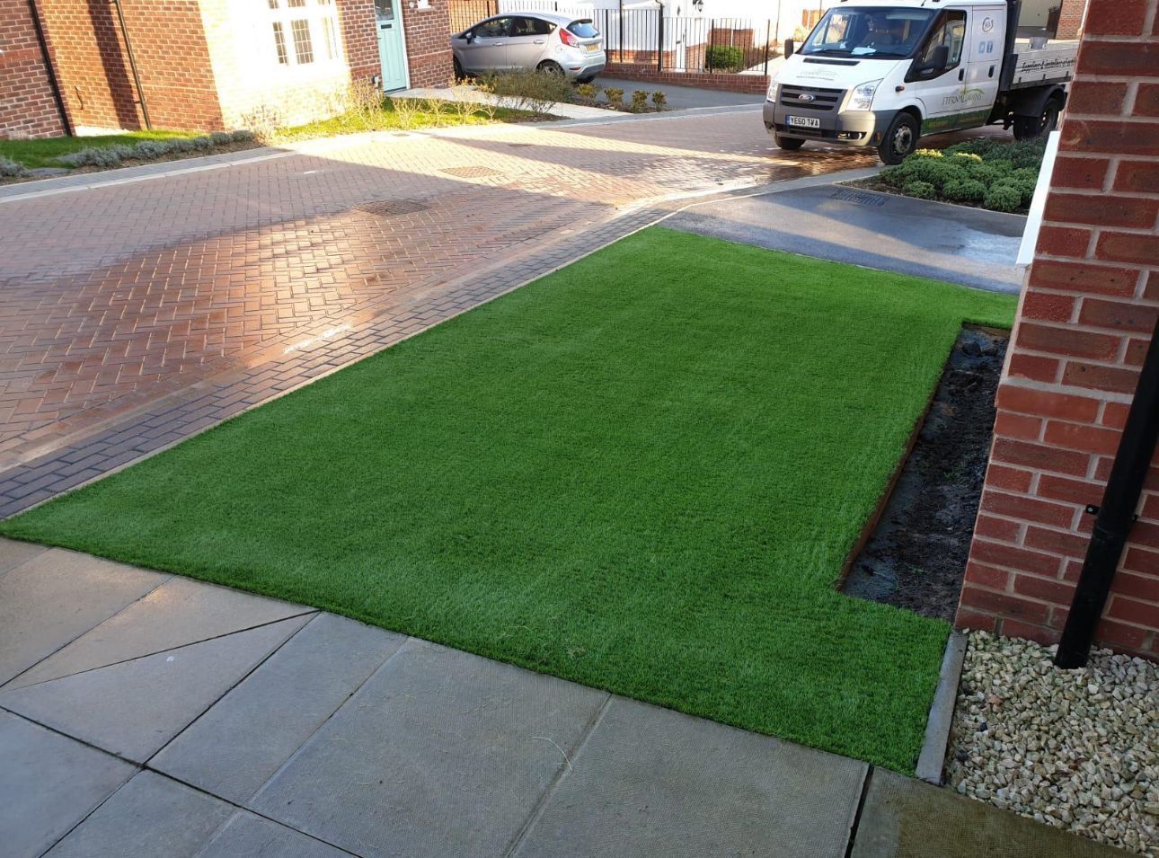 Front yard with fresh green turf bordered by paving stones and a driveway with parked cars.