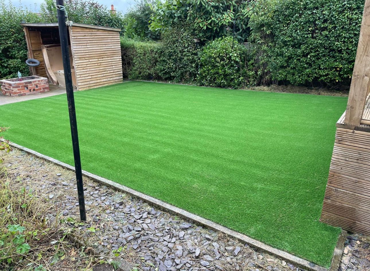 Backyard with bright green artificial turf, gravel border, and a small wooden shed beside hedges