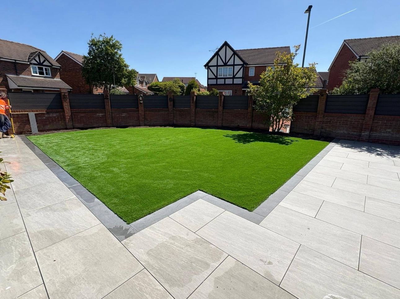 Backyard garden with green lawn, stone patio, and brick fence under a sunny sky