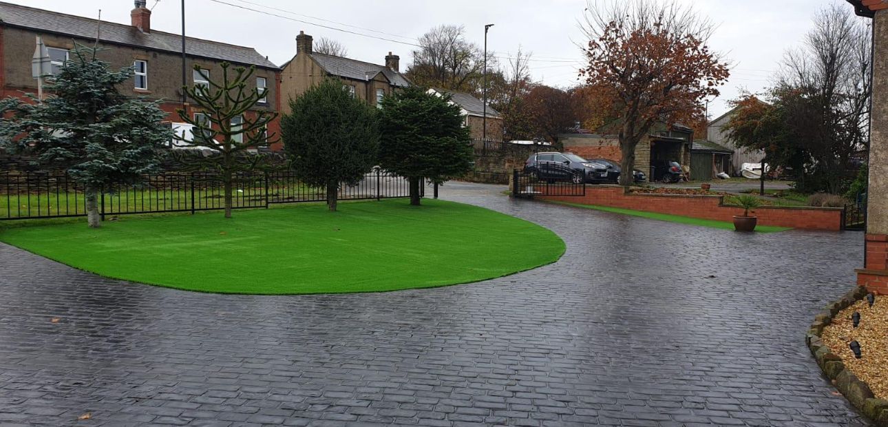 Rainy cobblestone courtyard with a bright green lawn, trees, and brick houses in the background