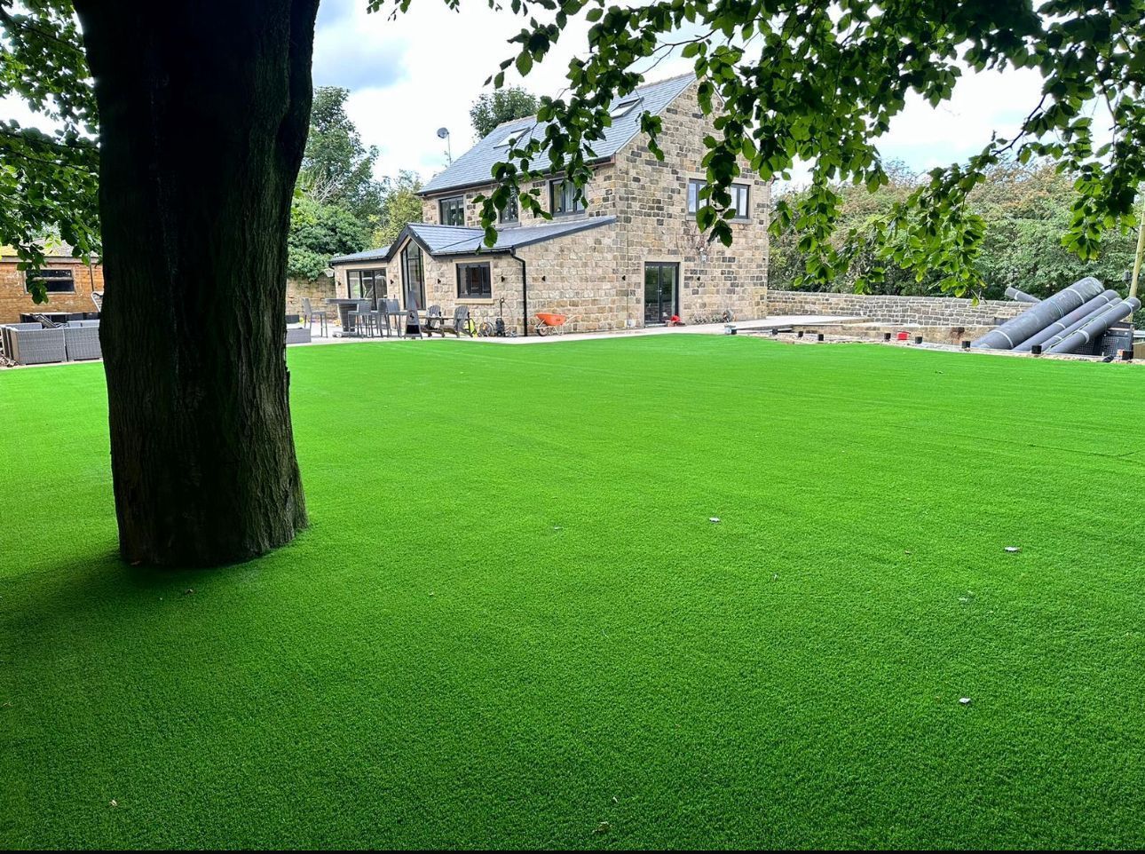Stone building beside a bright green lawn with a tree in the foreground and stone wall in back