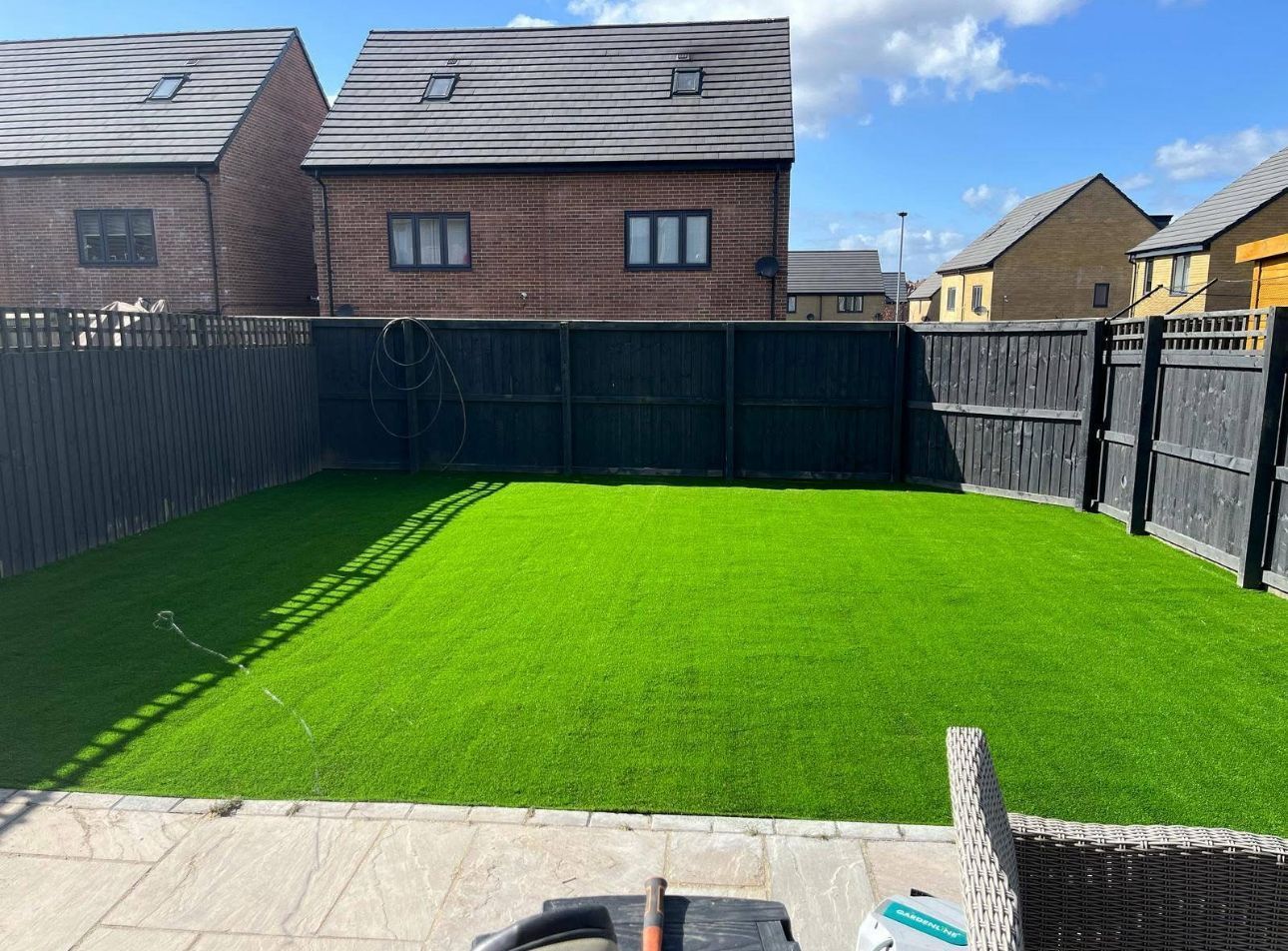 Backyard with bright green artificial lawn, dark fencing, and brick houses under a blue sky