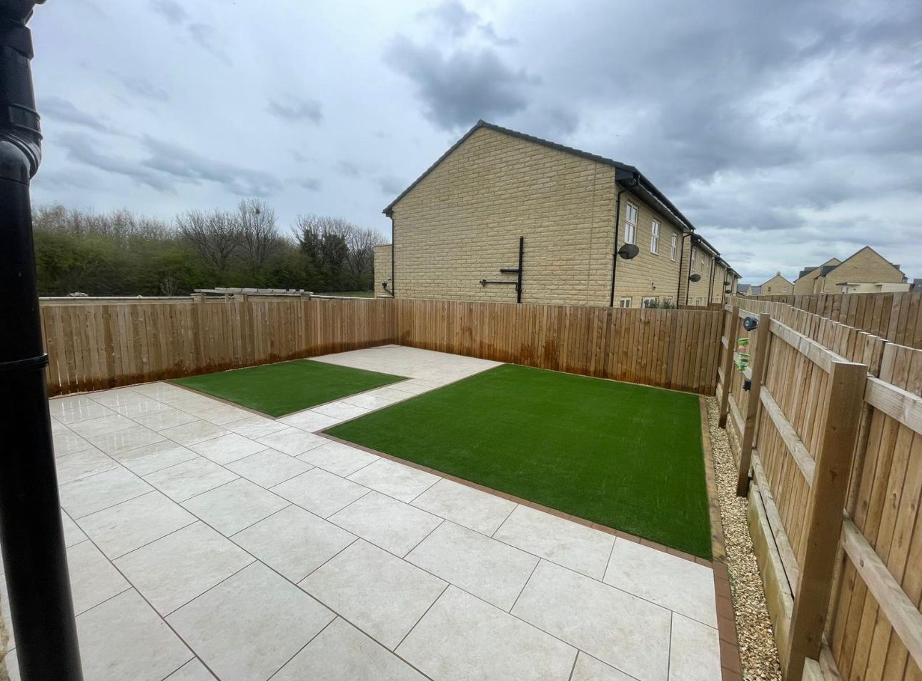 Backyard patio with gray pavers, artificial grass, and wooden fences under a cloudy sky