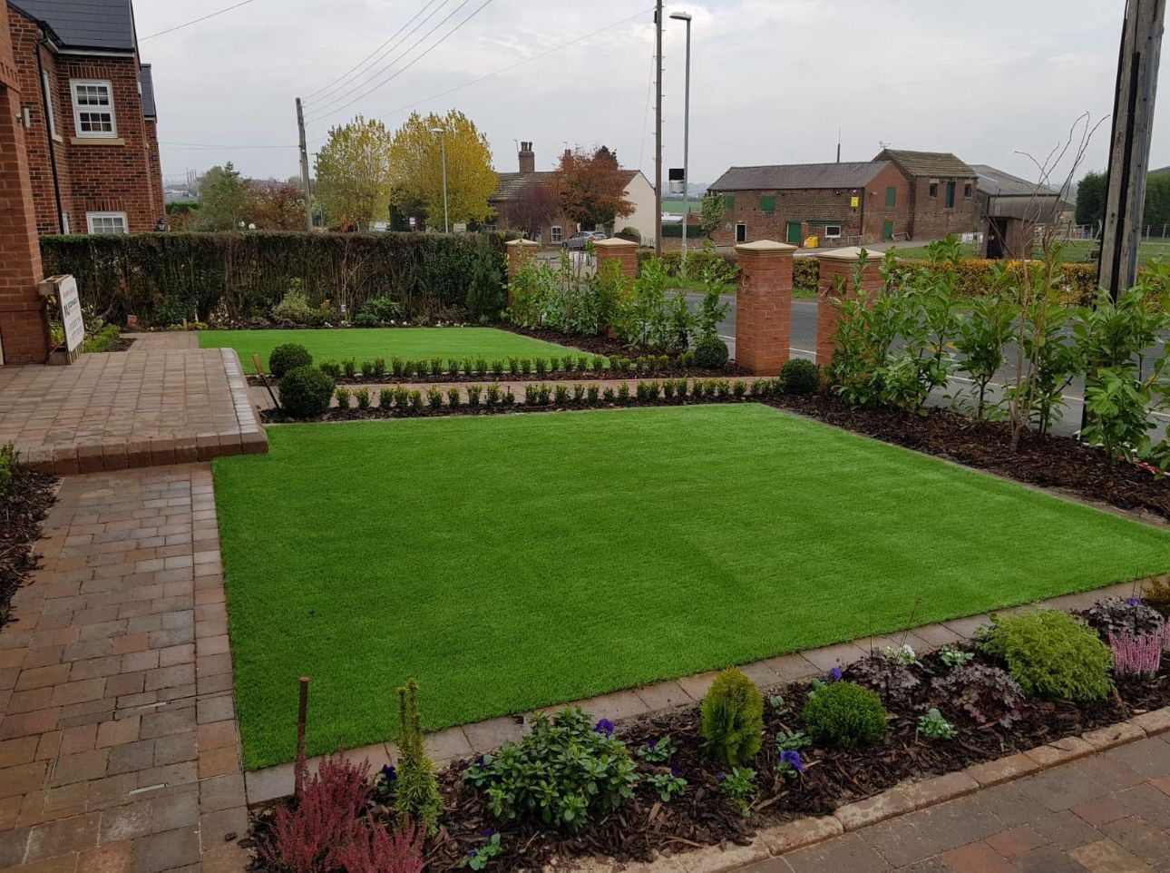 Small landscaped garden with bright green lawn, brick patio, flower beds, and houses in the background