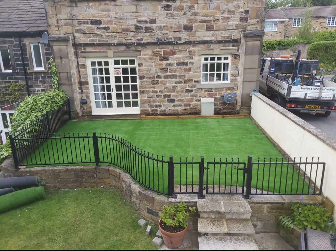Stone house backyard with bright green lawn, black metal fence, steps, and patio doors