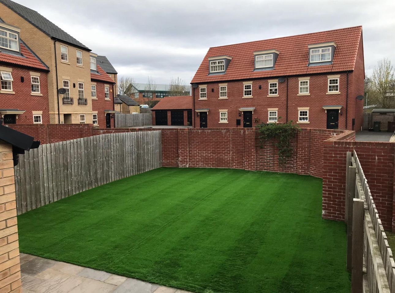Backyard with bright green lawn, brick patio, and red-roofed houses beyond the fence