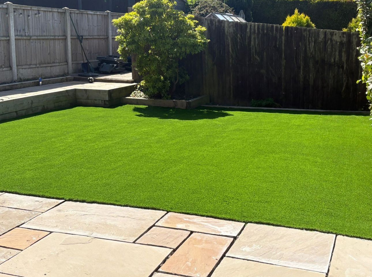 Backyard with bright green artificial grass, stone patio, wooden fence, and a leafy shrub.