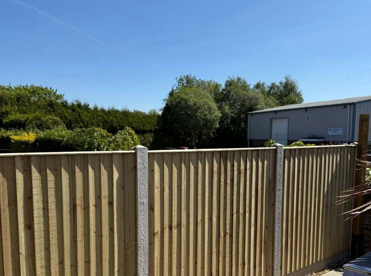 Wooden fence in front of green trees and a gray shed under a clear blue sky
