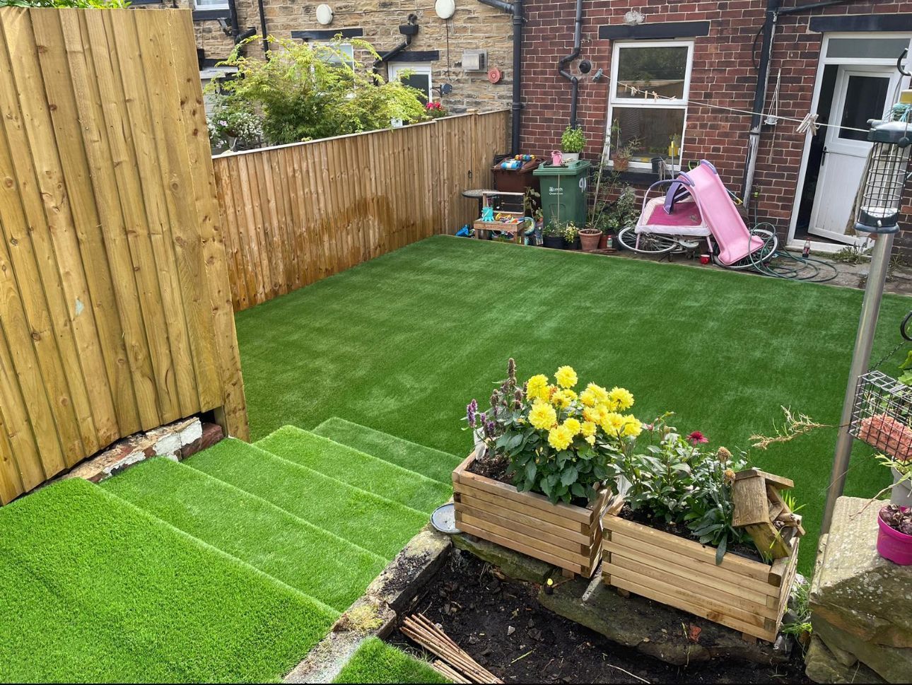Small backyard with artificial grass, wooden fence, flower bed, and pink children’s ride-on toy near a brick wall