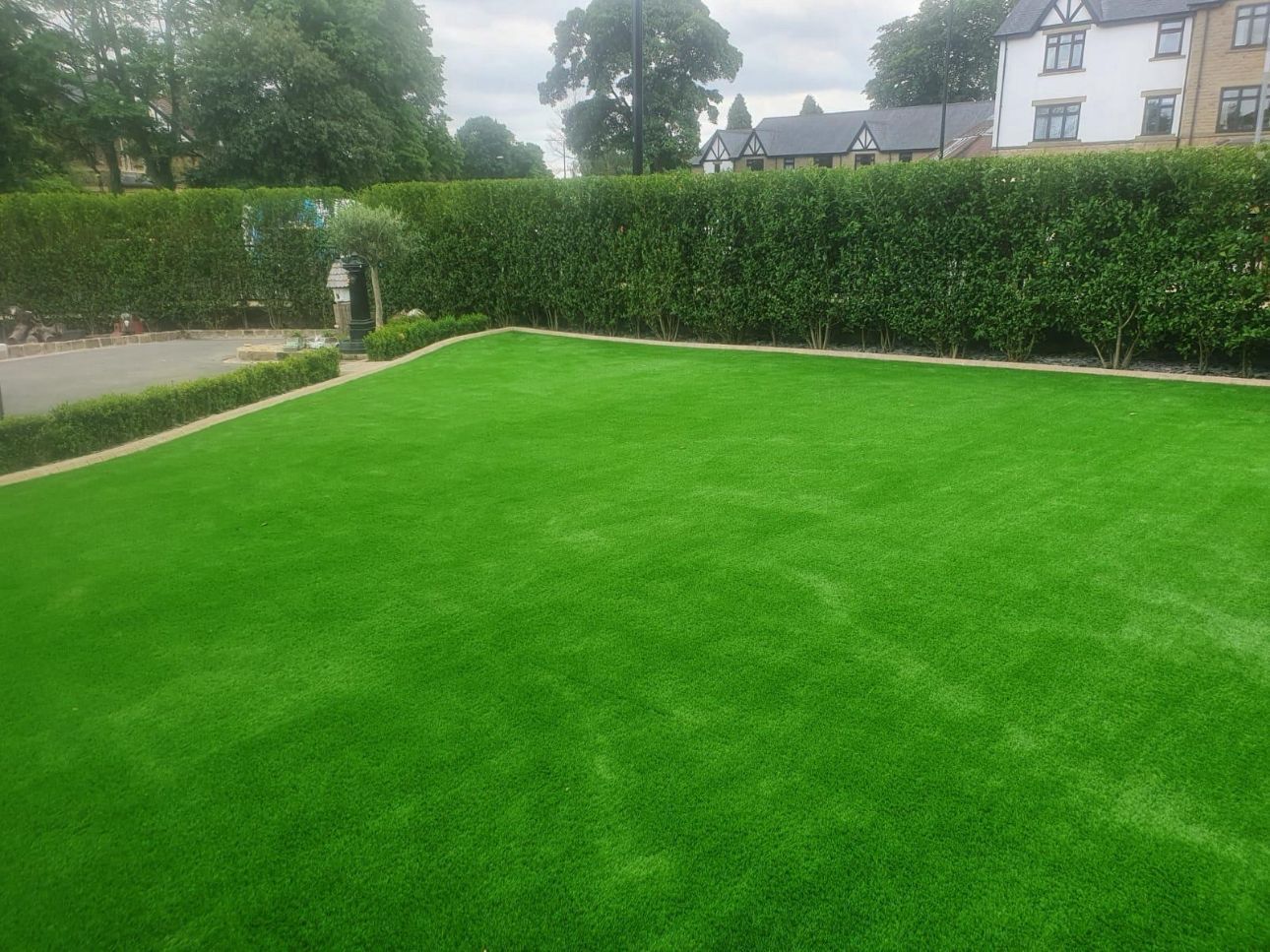 Well-kept green lawn bordered by hedges, with houses and trees in the background.