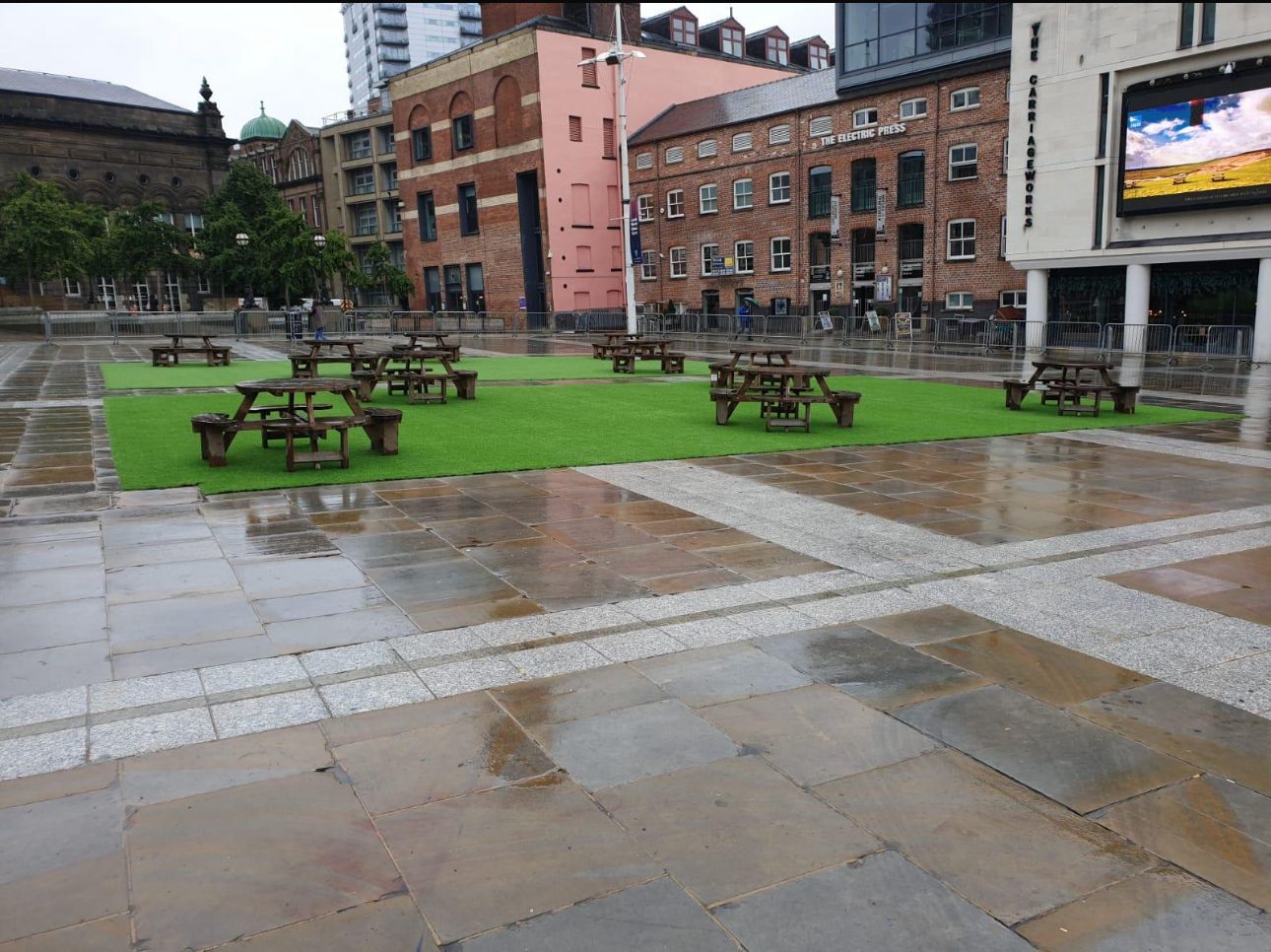 Rainy urban plaza with picnic tables, wet stone paving, grass, and brick buildings in the background