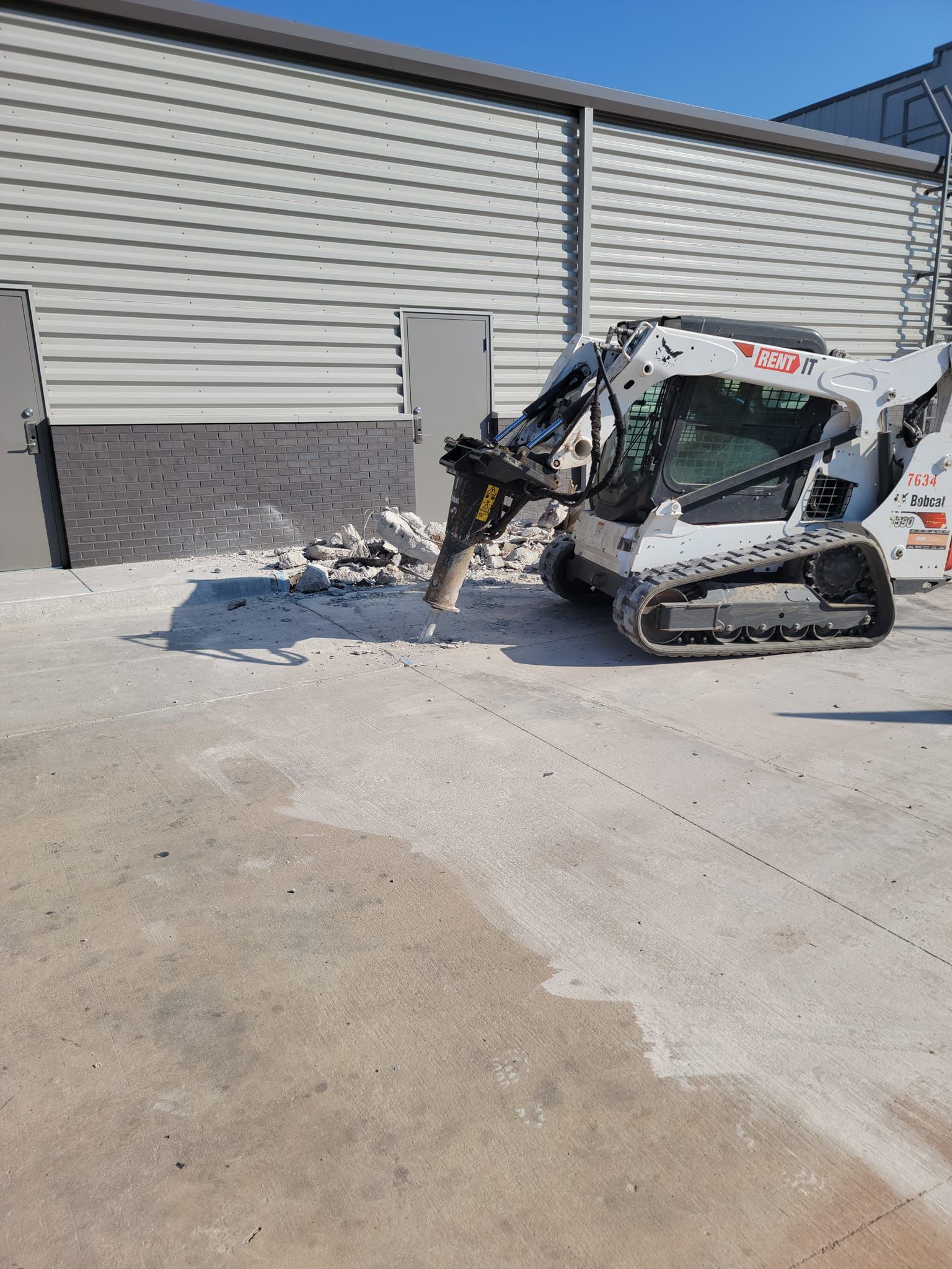 White skid-steer loader parked beside a gray building on a concrete lot, with debris near the wall.