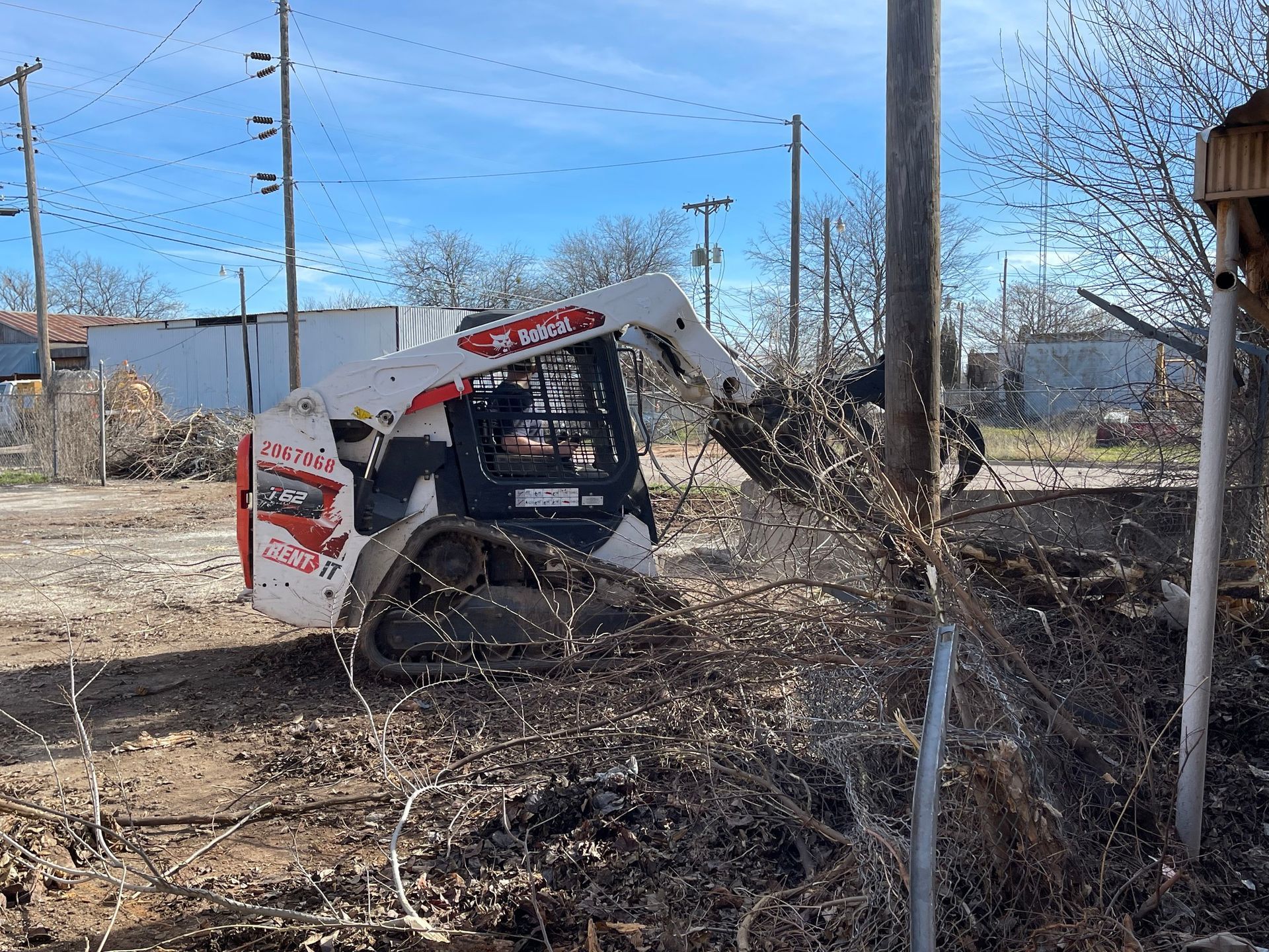 Bobcat skid-steer stuck in brush beside a chain-link fence and utility pole on a clear day