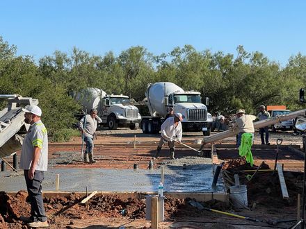 Construction workers pouring concrete at a building site with trucks and trees in the background
