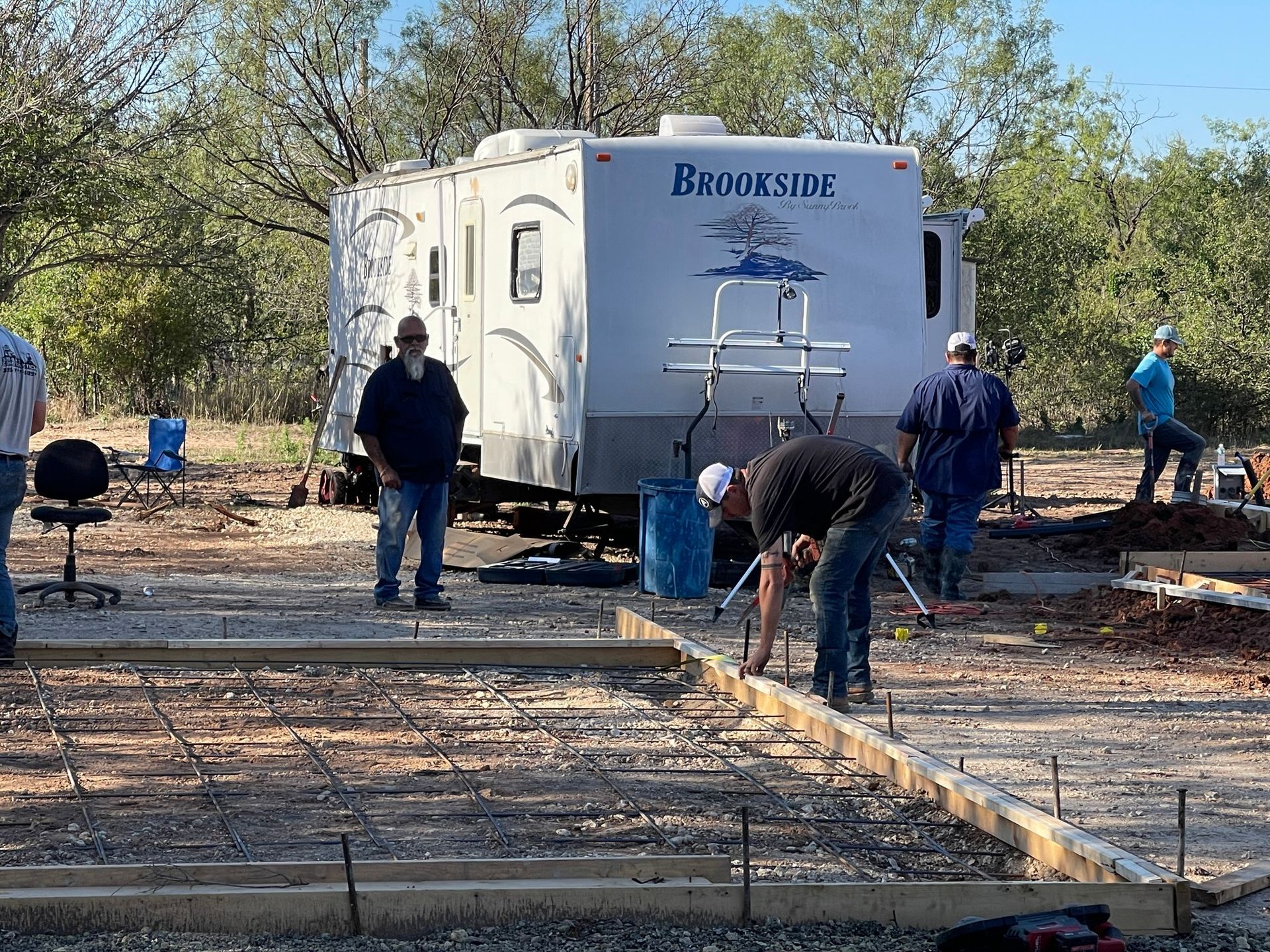 Workers near a white trailer at a debris-filled construction site, with concrete forms in the foreground