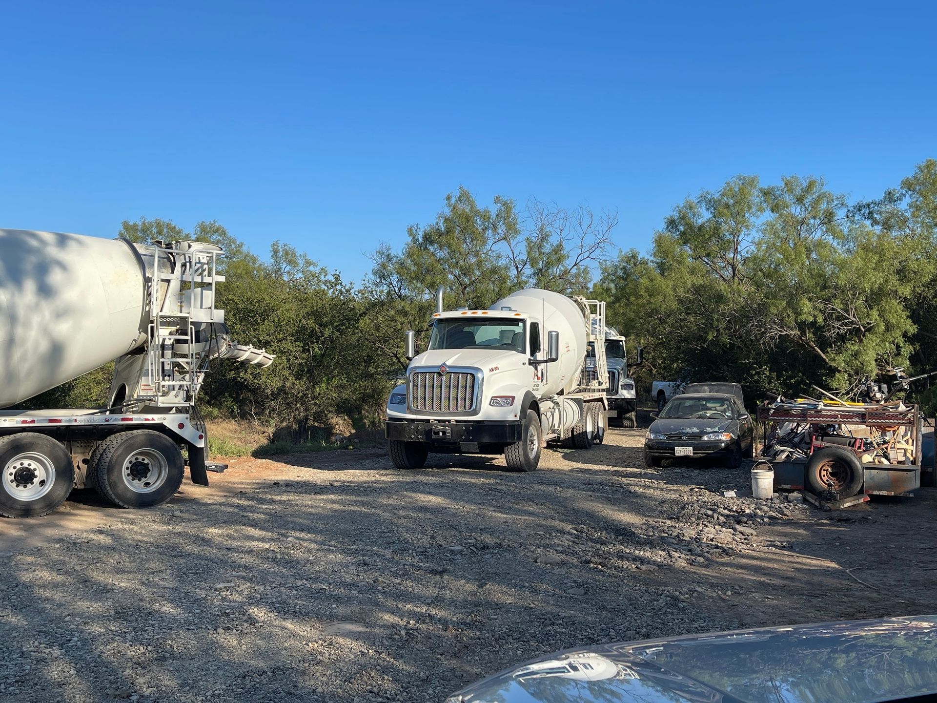Construction vehicles parked on a gravel lot beside trees under a blue sky