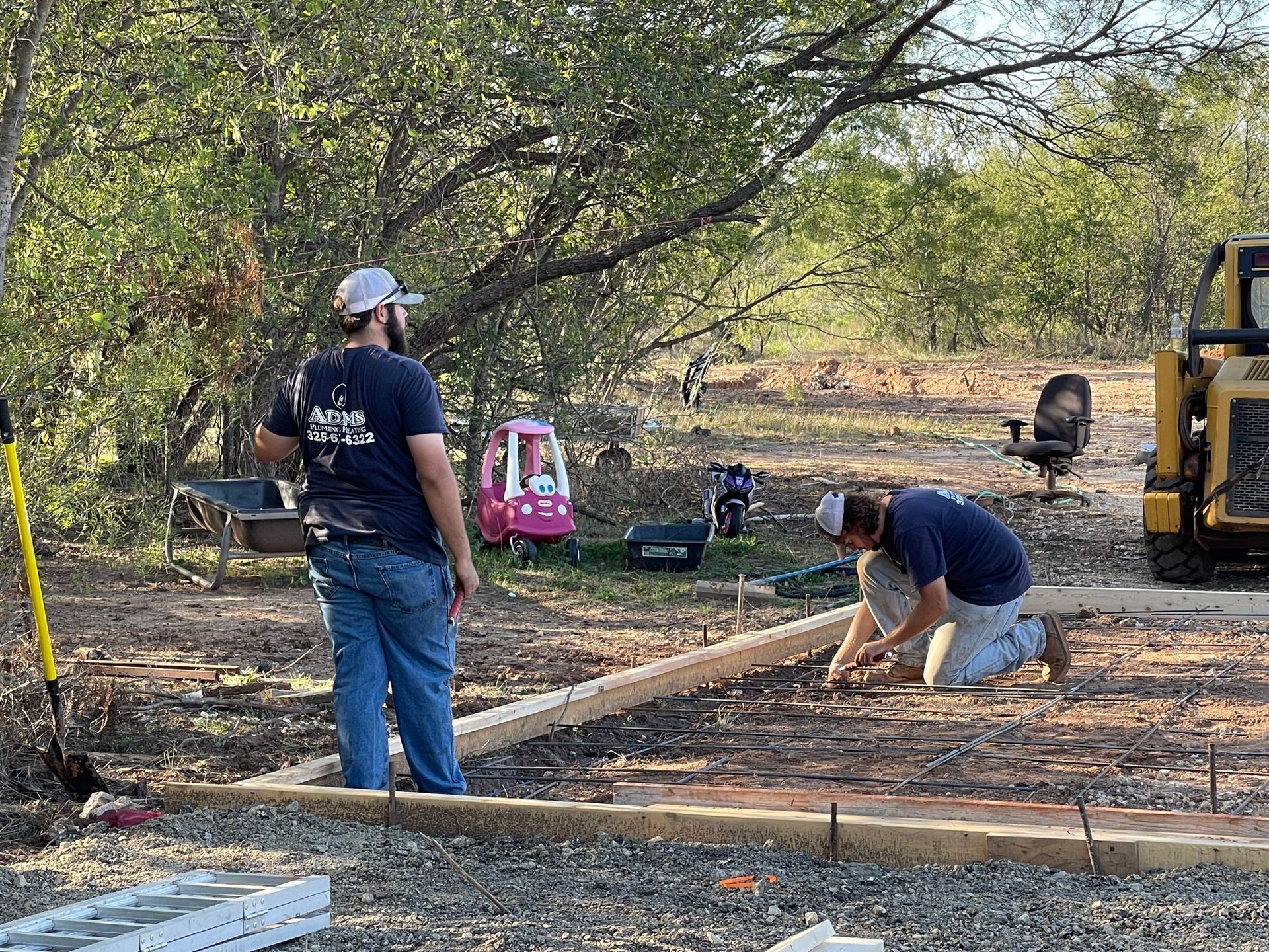 Two workers building a wooden frame outdoors among trees and dirt, with a pink tool bag nearby.