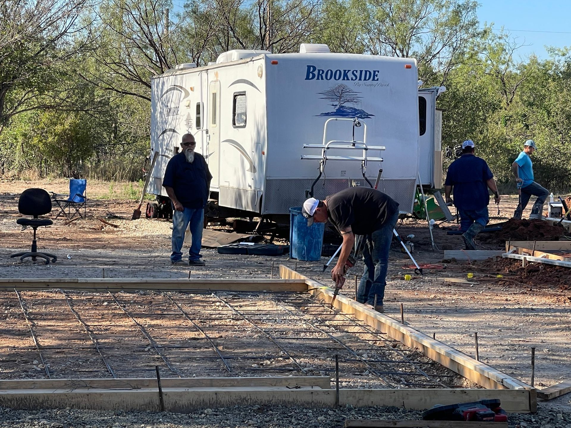 People working on a concrete slab foundation near a Brookstone trailer in a wooded area.