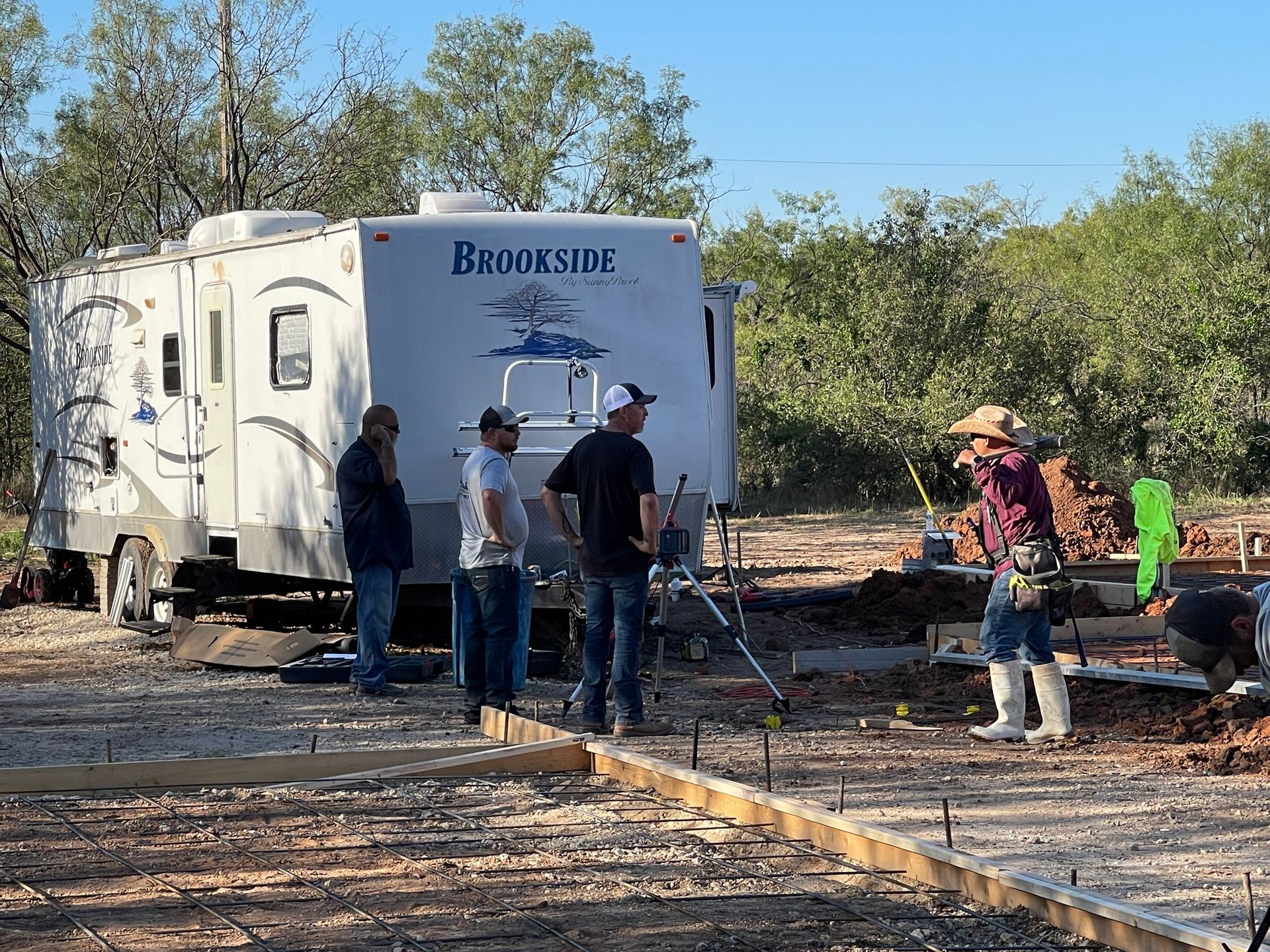 Workers beside a Brookside trailer on a dirt worksite, with tools and materials laid out nearby.