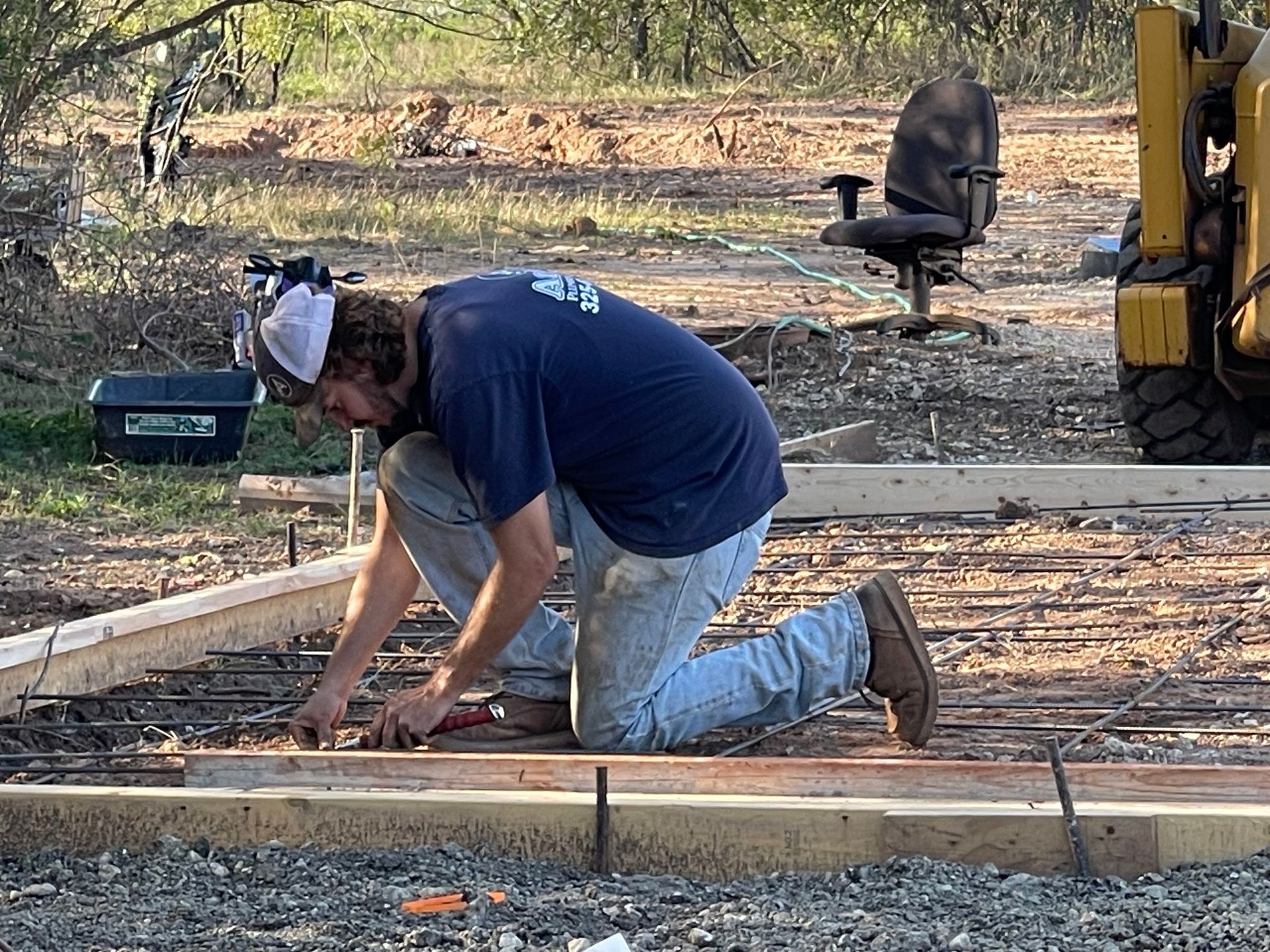 Worker kneeling on dirt, measuring and marking a wooden form at a construction site.
