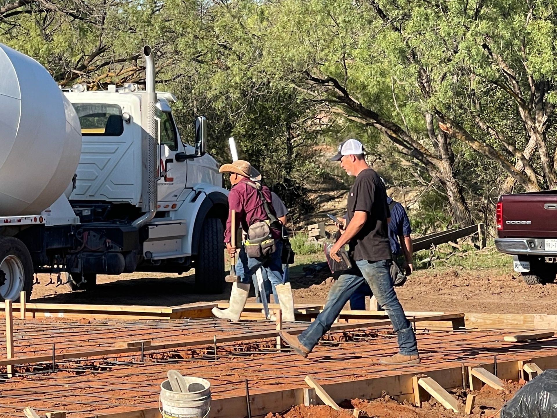 Workers walking beside a concrete truck at a construction site with lumber and trees nearby