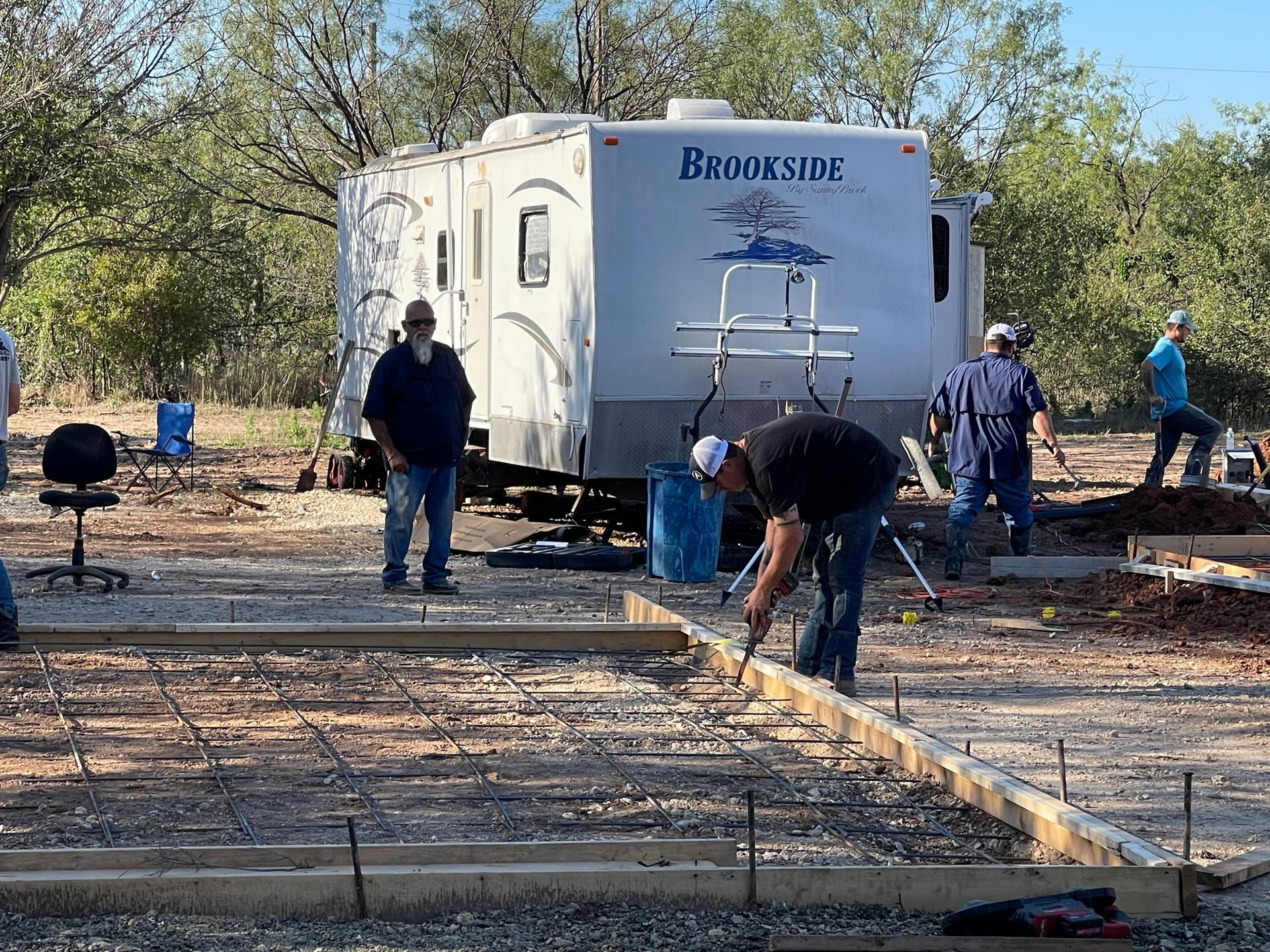 Workers building a foundation beside a white trailer in a wooded outdoor area