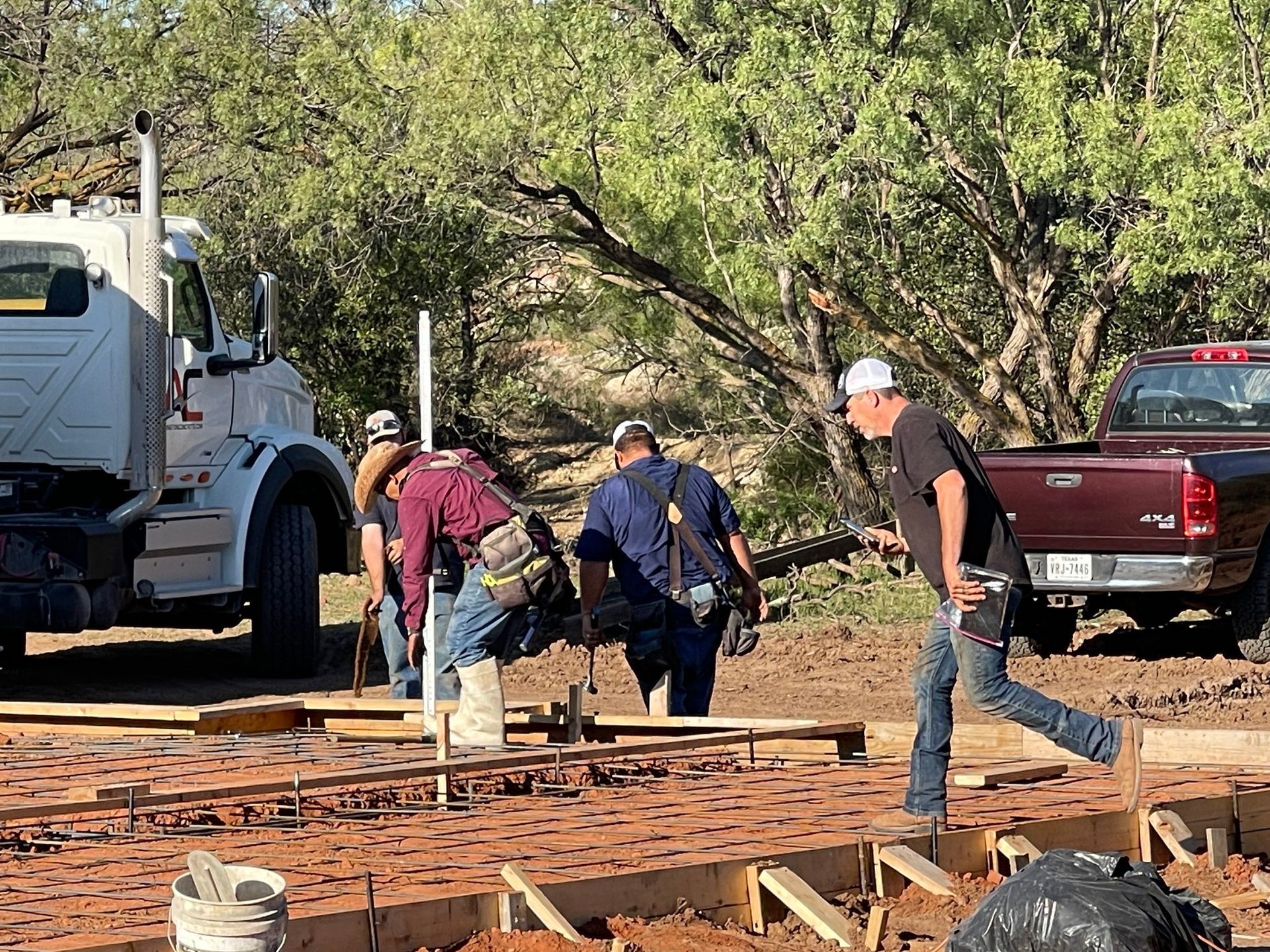 Workers building a wooden foundation beside a white truck and red pickup in a wooded outdoor area
