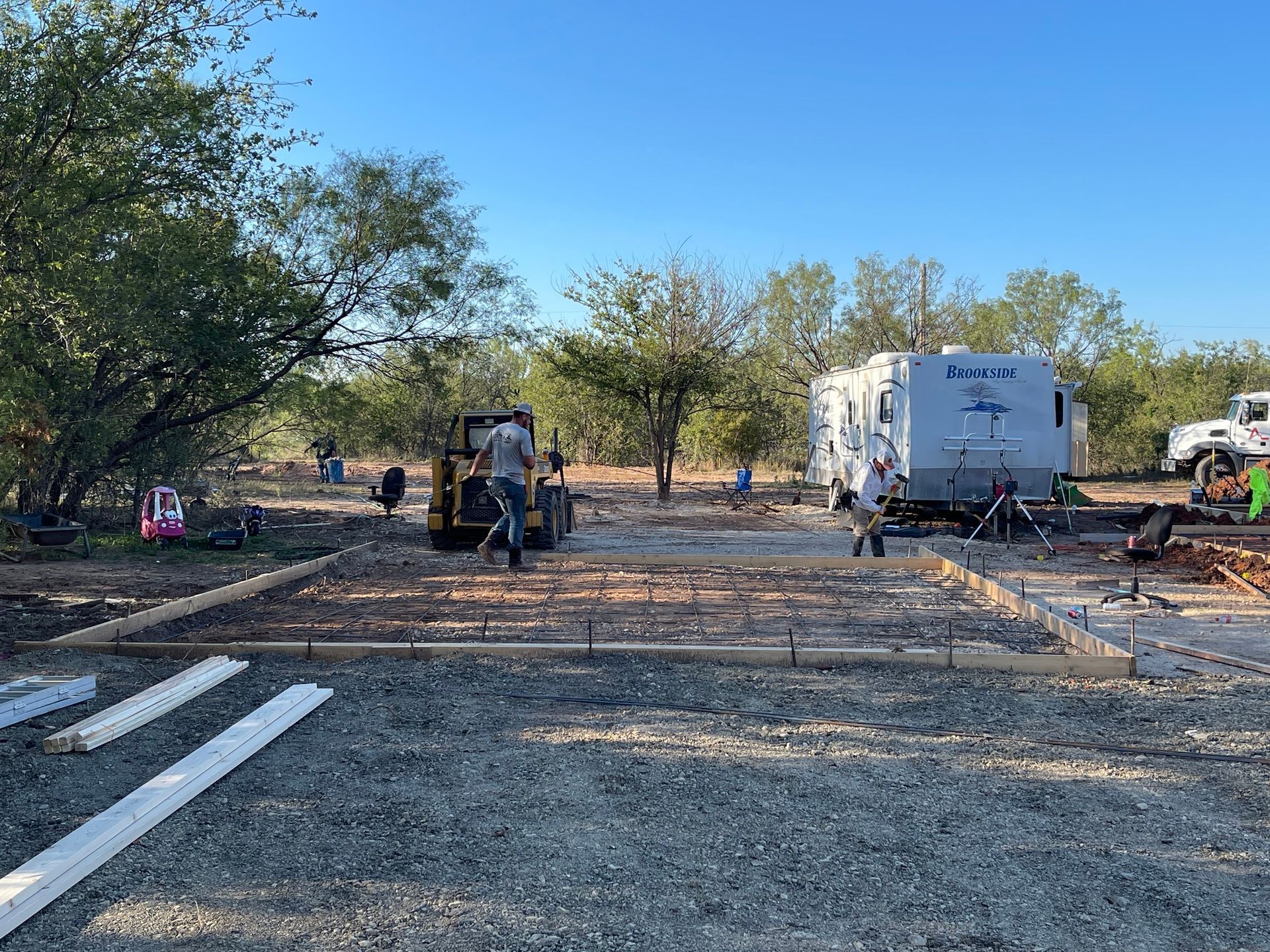 Construction site with excavator, white truck, and workers on a gravel lot under trees