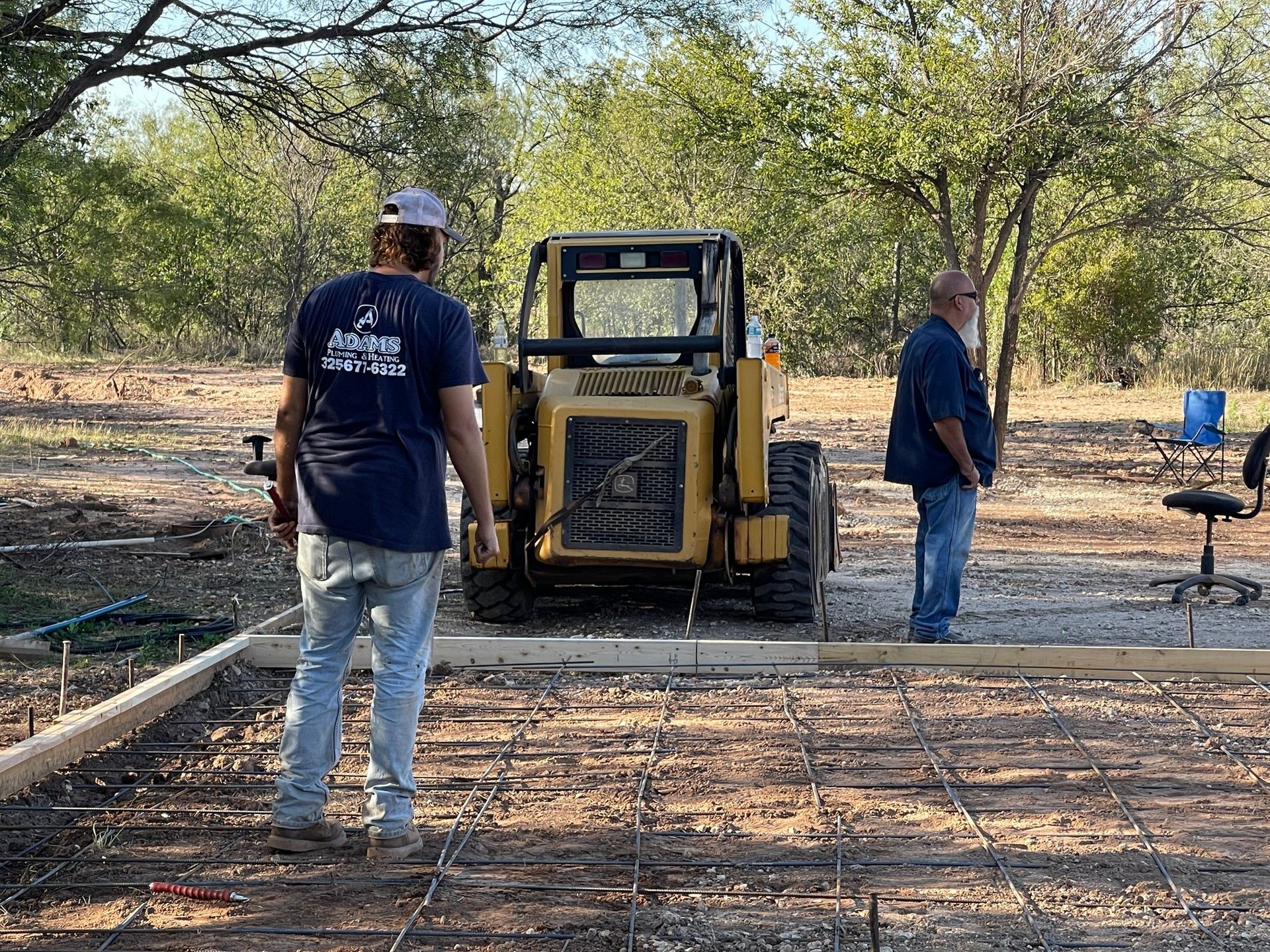 Workers stand near a yellow skid steer on a dirt lot with trees and pipes in the background.