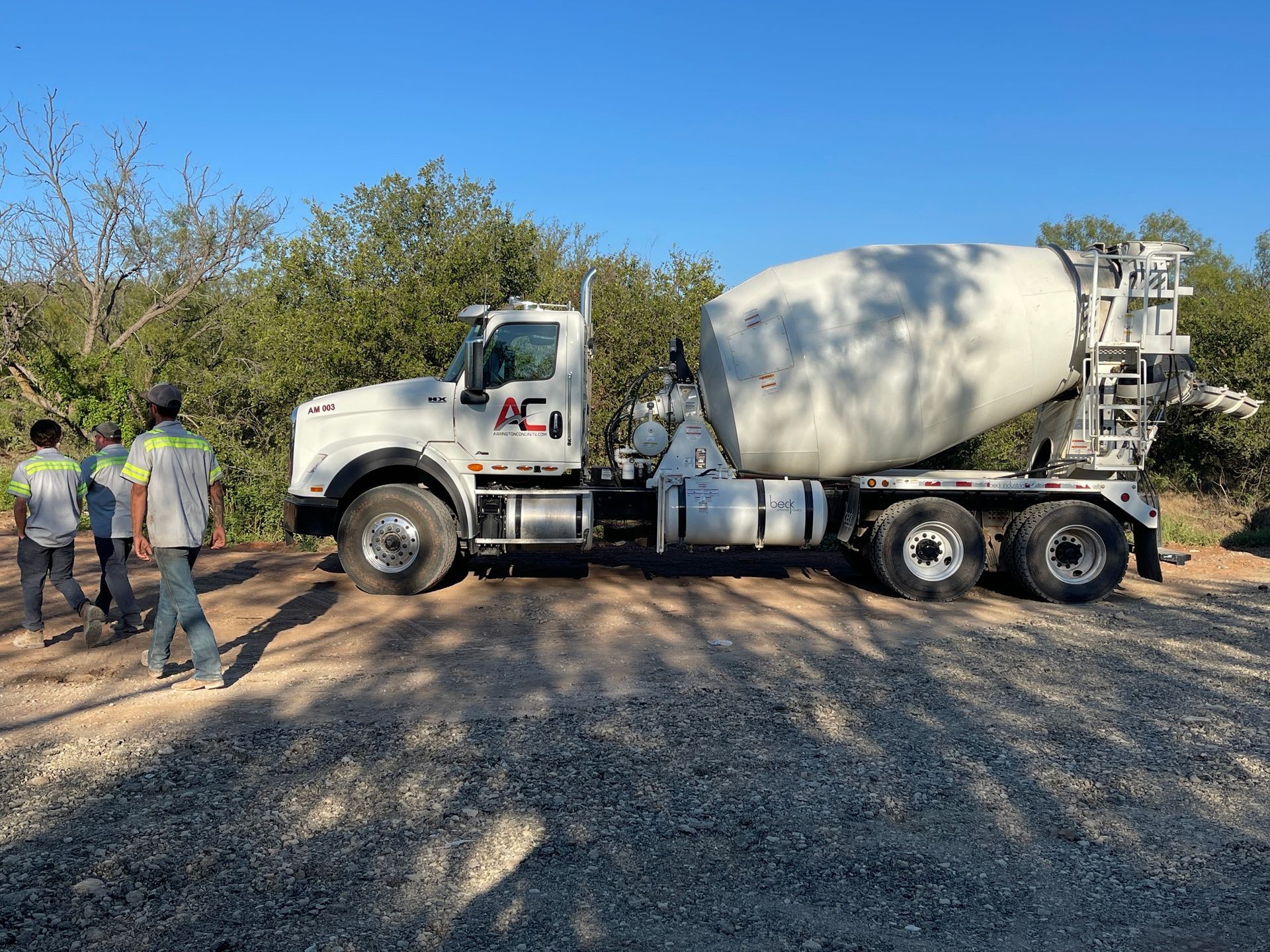 Cement mixer truck parked on a gravel road with three workers standing beside it under trees.