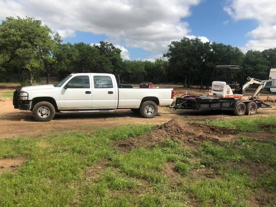 White pickup truck towing a black trailer with equipment on a dirt path in a grassy rural area