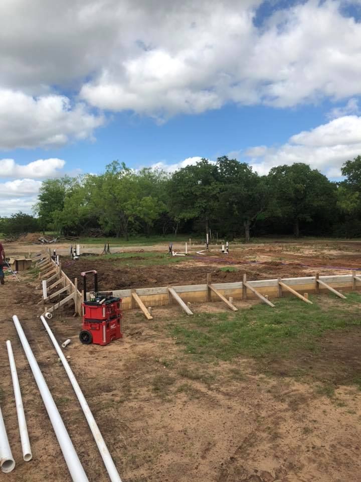 Construction site with wooden foundation forms, white pipes, and red equipment under a cloudy sky