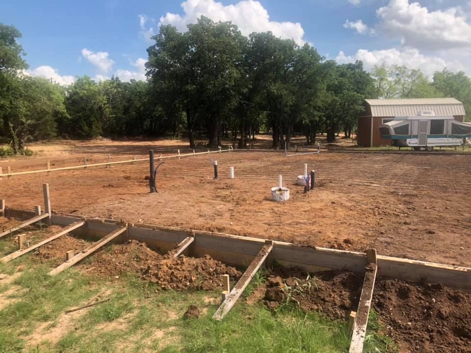 Dry dirt lot with trees and a small shed, with wooden boards and concrete forms in the foreground.
