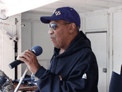 Man speaking into a microphone, wearing a blue cap and hoodie. Outdoors setting.
