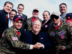 Group of men, some in camouflage, pose for a photo. Some wear baseball caps, smiles, and handshakes.