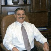 Man smiling in a white shirt and tie, sitting in a leather chair in an office setting.
