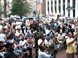 Large outdoor gathering clapping, in front of buildings. Some people are seated. FedEx sign in background.