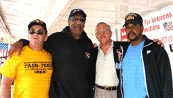 Four men in casual attire pose, arms around each other. One wears a yellow shirt, others hats. Signs with