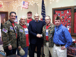 Group of men, some in camouflage shirts, with a man in a suit, posing indoors near an American flag.