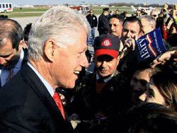 Bill Clinton smiles and shakes hands with a crowd at an airport.