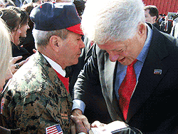 Bill Clinton shaking hands with a man in military fatigues and hat outdoors.
