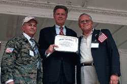 Three men on a stage; one in suit holds a certificate, flanked by men in military and patriotic attire.