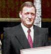 Man in suit, red tie, holding a framed document, smiling.