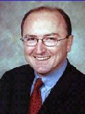 Man in glasses, smiling, wearing a suit and red tie, against a blurred background.