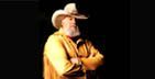 Man with white beard, cowboy hat, yellow jacket, arms crossed, on stage.