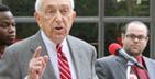 Man in gray suit speaks, pointing; two men stand behind him.