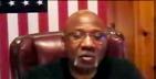 Man with glasses speaks, seated in front of an American flag.