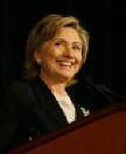 Hillary Clinton smiling at a podium, wearing a dark suit jacket, white blouse.