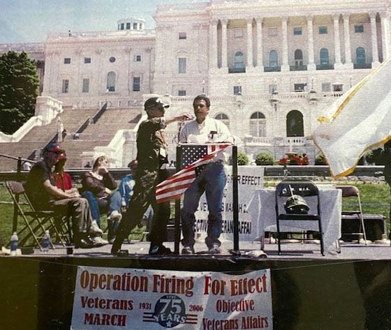 Veterans protest at the US Capitol. A man hands a flag to a soldier on a stage with a banner: